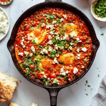 Shakshuka with feta and chorizo in a skillet with bread, eggs and napkin around the pan.