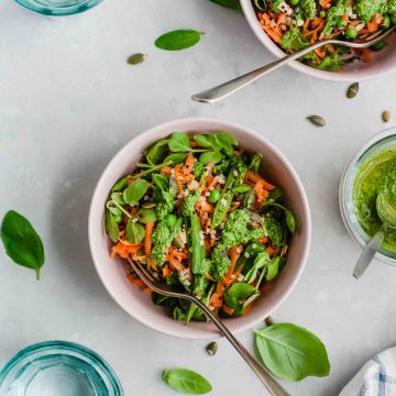 Two bowls of quinoa salad with pesto and peas and forks, with basil leaves nearby.