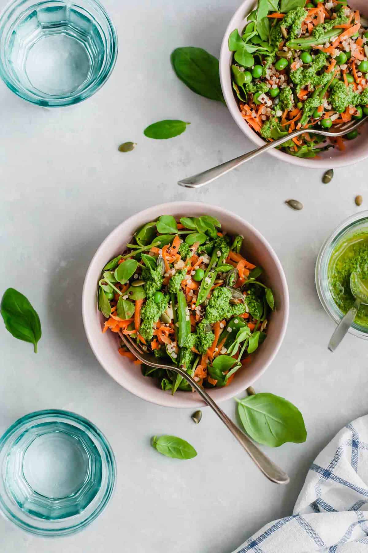 Two bowls of quinoa salad with pesto and peas and forks, with basil leaves nearby.