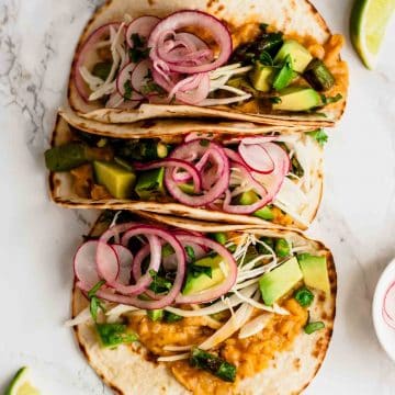 An array of refried bean tacos with avocado and pickled onions for toppings.