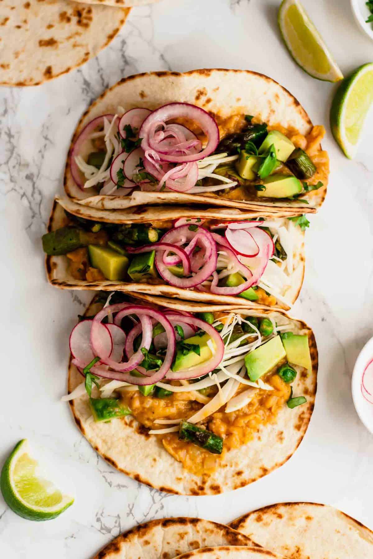 An array of refried bean tacos with avocado and pickled onions for toppings. 