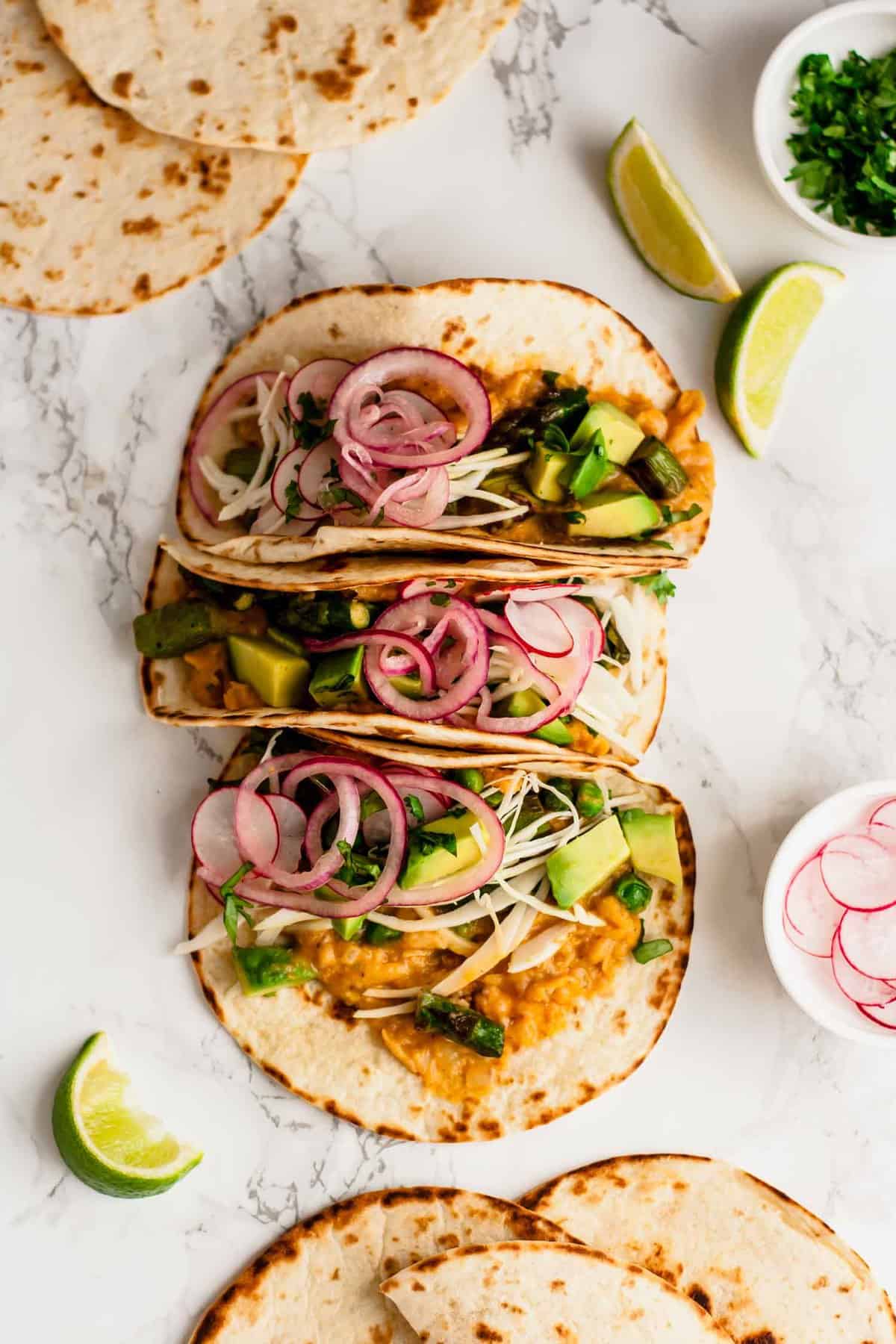 Three vegan tacos with refried beans and asparagus with pickled onions on top.
