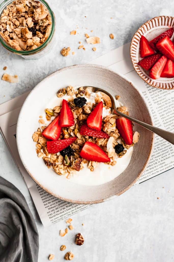 coconut tahini pistachio granola in a bowl with yoghurt and strawberries with a spoon and granola scattered around