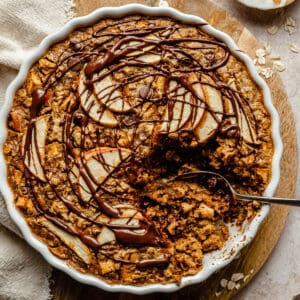 Apple baked oatmeal in a baking dish being served up with a spoon on a wooden board.