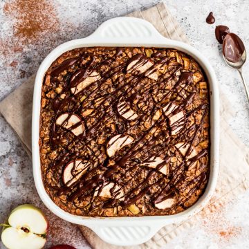 Apple baked oatmeal in a baking dish with apples to the side and drizzled with dark chocolate.