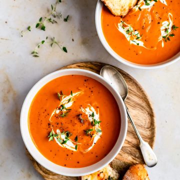 Two bowls of butternut squash and red pepper soup one on a board with bread and a spoon.