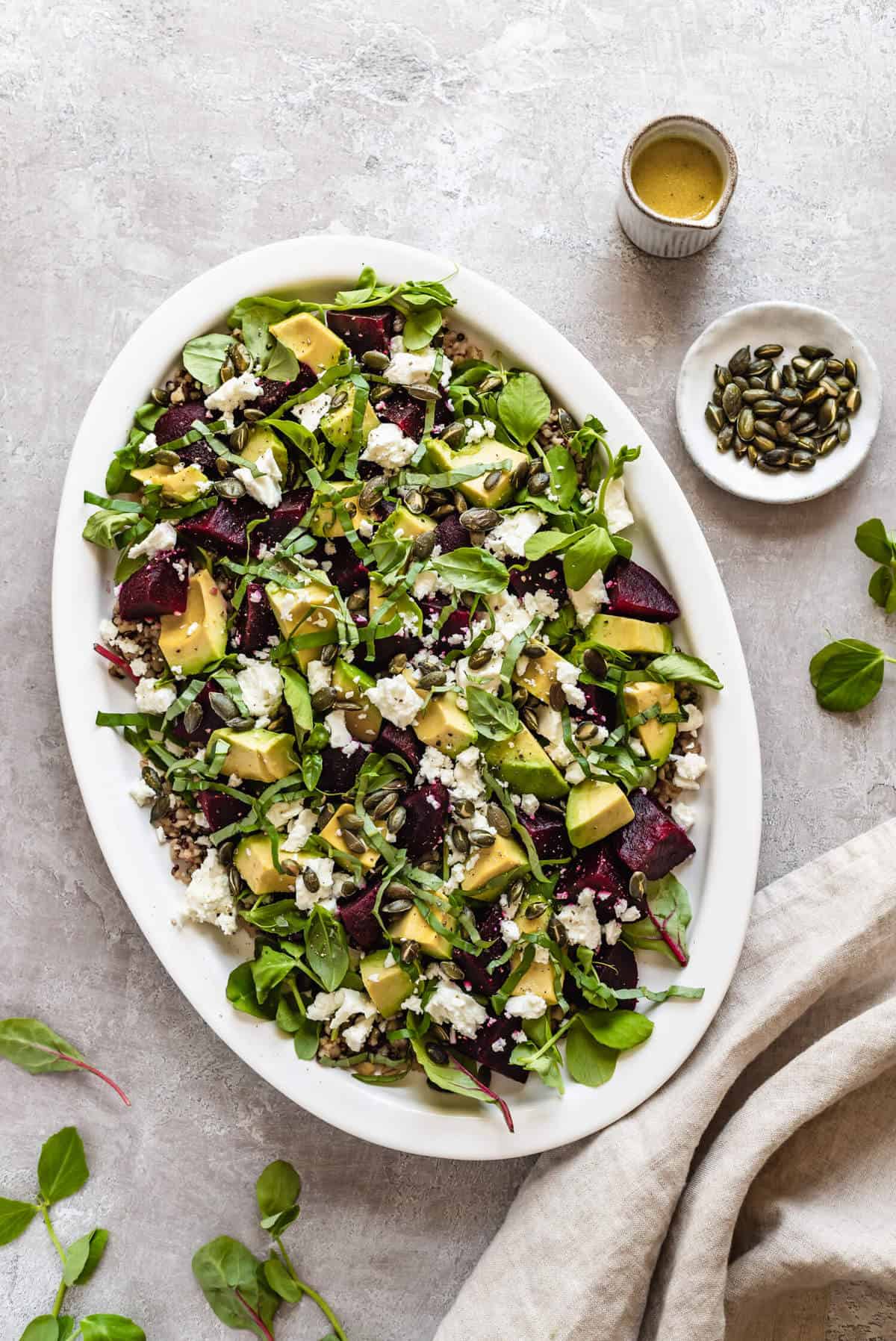 a platter of beetroot avocado feta salad on a light background with a small jug, beige napkin, pumpkin seeds and salad leaves around the outside