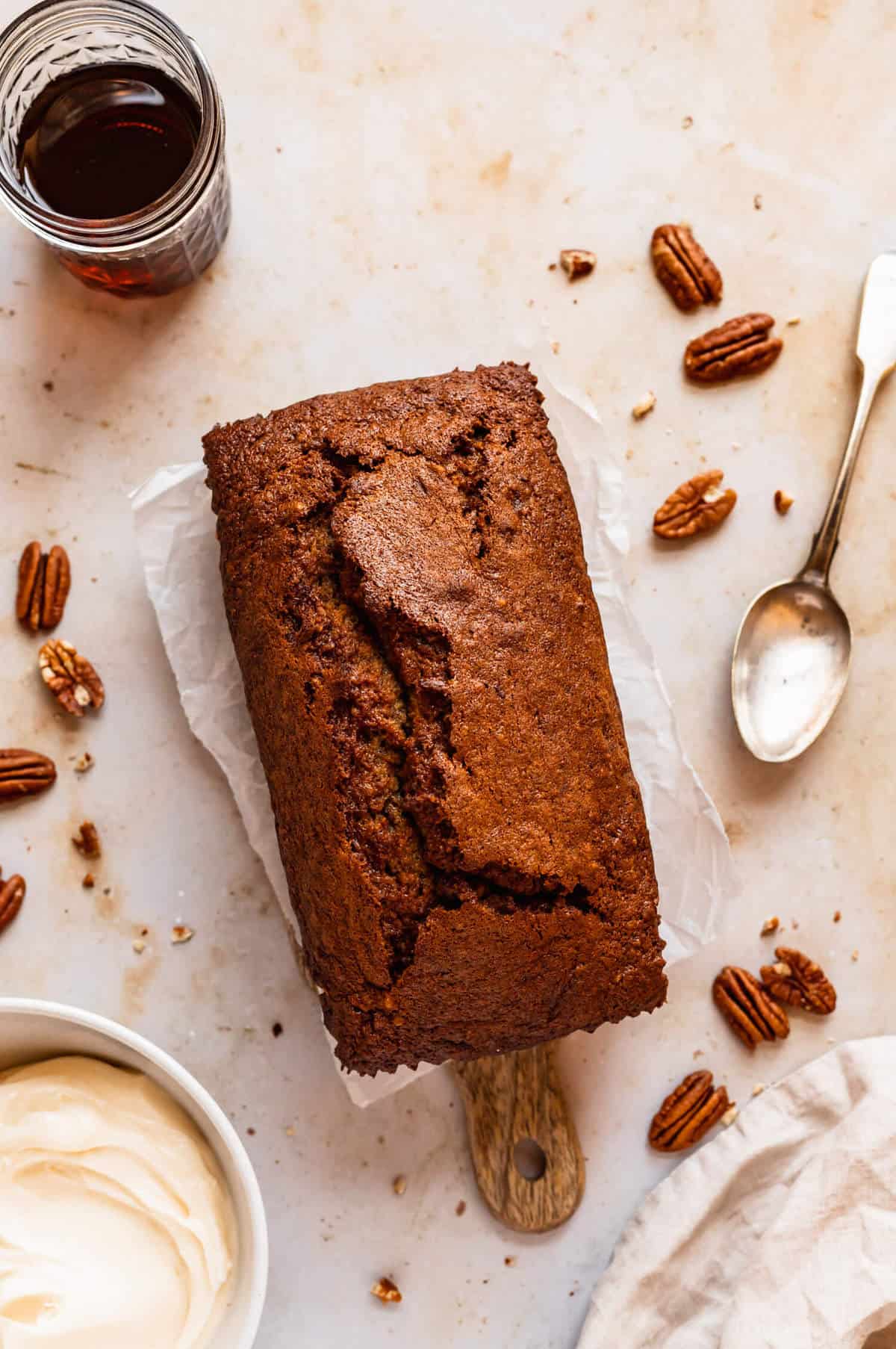 a loaf of banana bread surrounded by pecans, a jar of maple syrup and a bowl of frosting