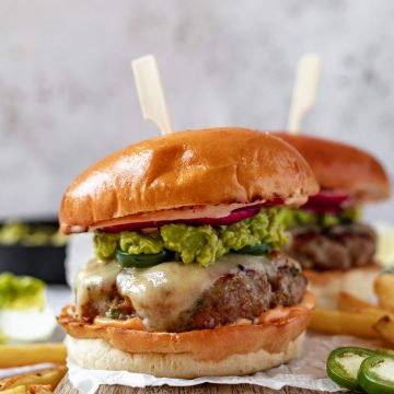 a close up of a burger on a wooden board covered in melted cheese and smashed avocado with a skewer in the top