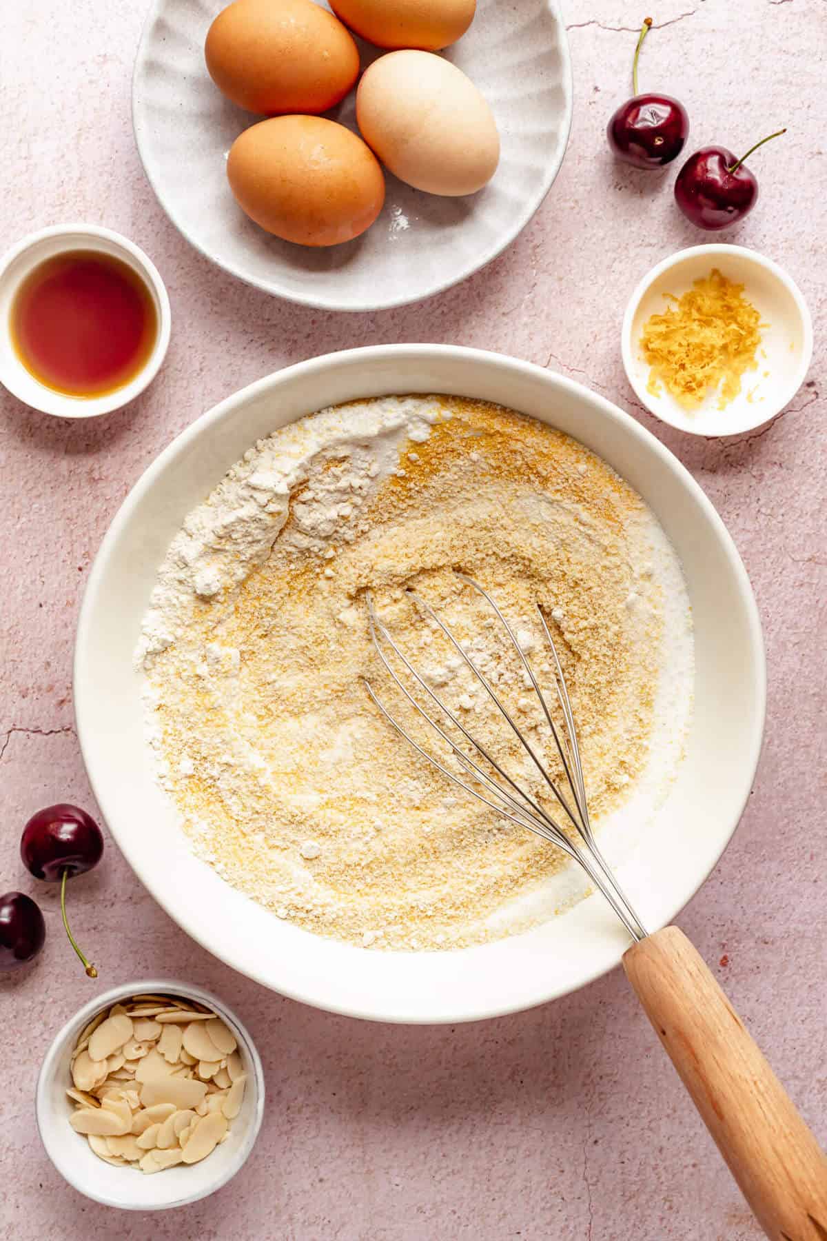 a mixing bowl filled with dry ingredients and a whisk, surrounded by cherries, eggs and flaked almonds