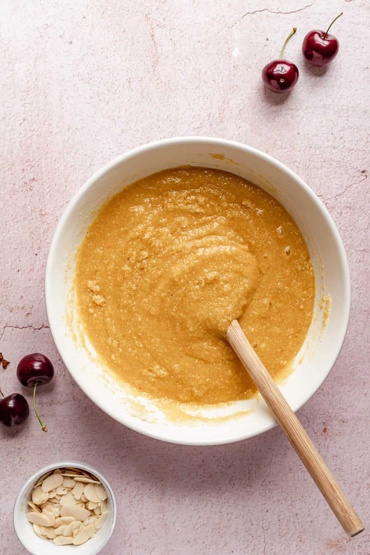 a bowl of cake batter with a spoon in it with some cherries and a bowl of flaked almonds on the side