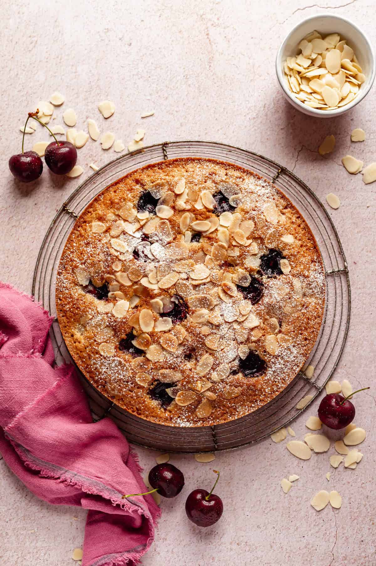 a cake studded with cherries and topped with flaked almonds on a round wire rack, surrounded by a pink napkin, cherries and flaked almonds