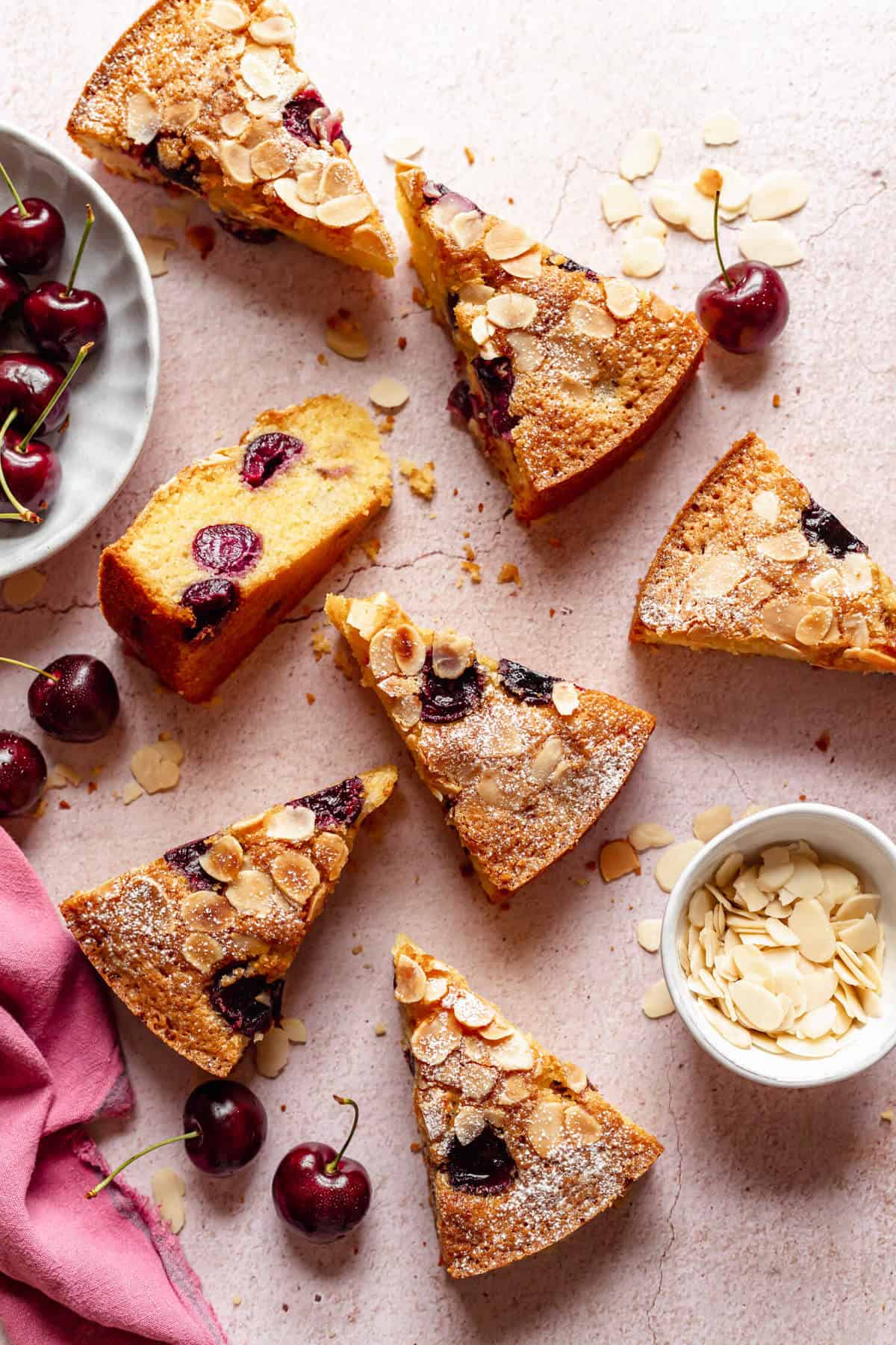 several slices of cake arranged on a peach plaster background, with a small bowl of flaked almonds and cherries