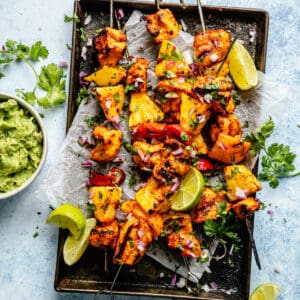 Grilled pineapple chicken kabobs served on a baking sheet with guacamole and lime wedges.