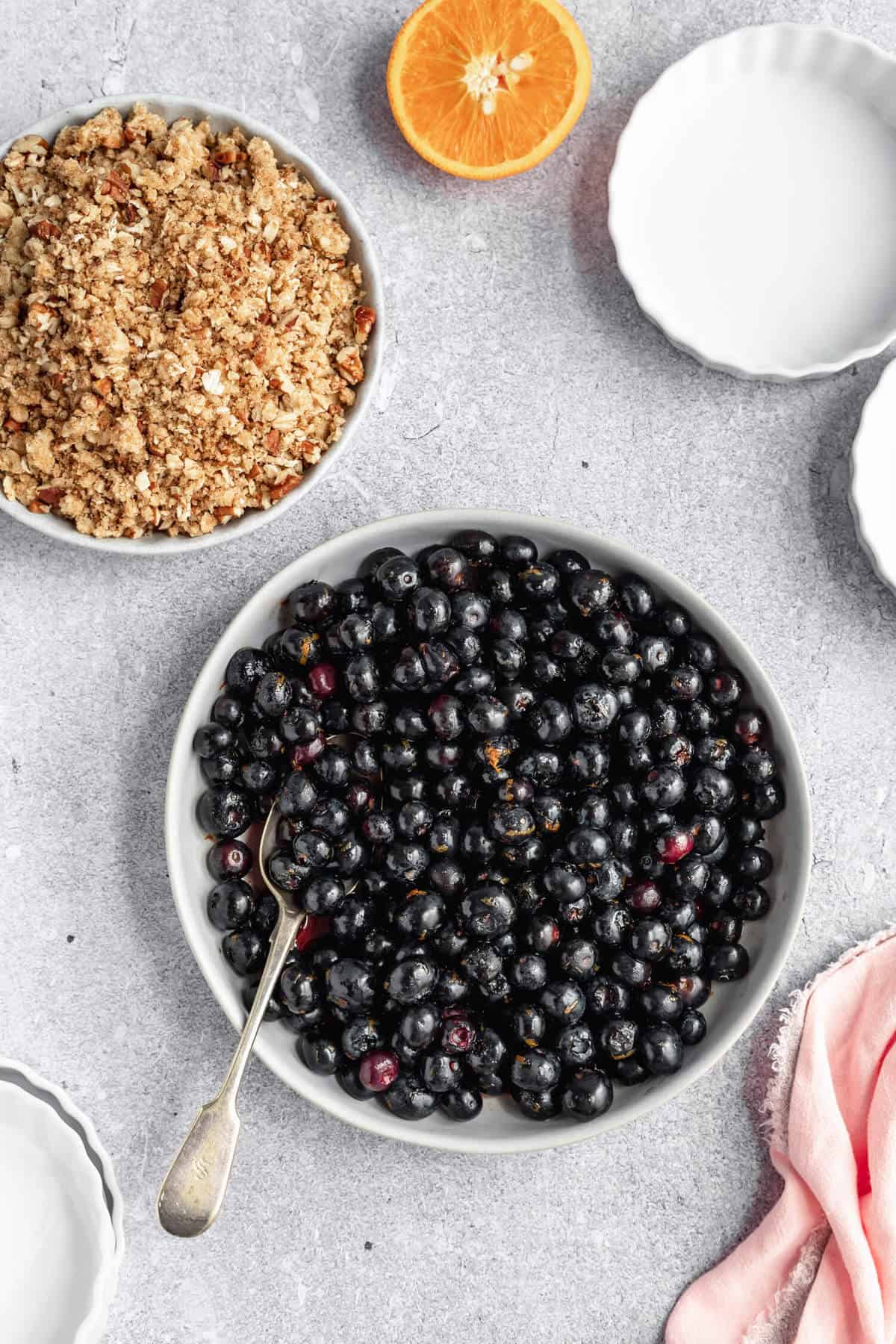 a bowlful of blueberries with a spoon, a bowl of crumble topping and half of an orange