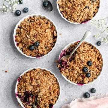 four ramekins filled with fruit crisp with spoons digging in and blueberries scattered around