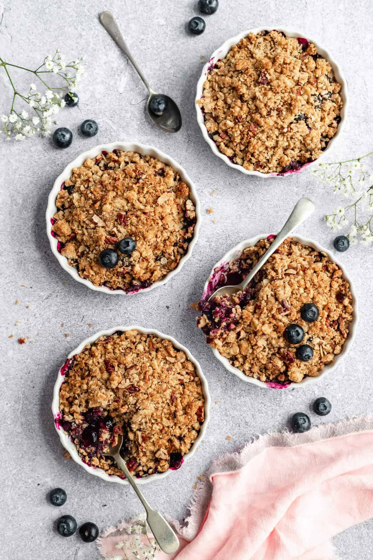 four ramekins filled with fruit crisp with spoons digging in and blueberries scattered around