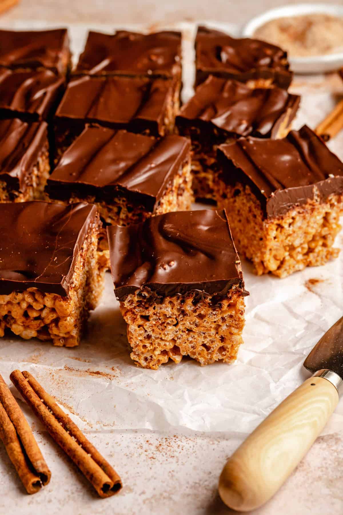 Rice krispie treats cut up into squares on parchment paper with a knife and cinnamon sticks in foreground.