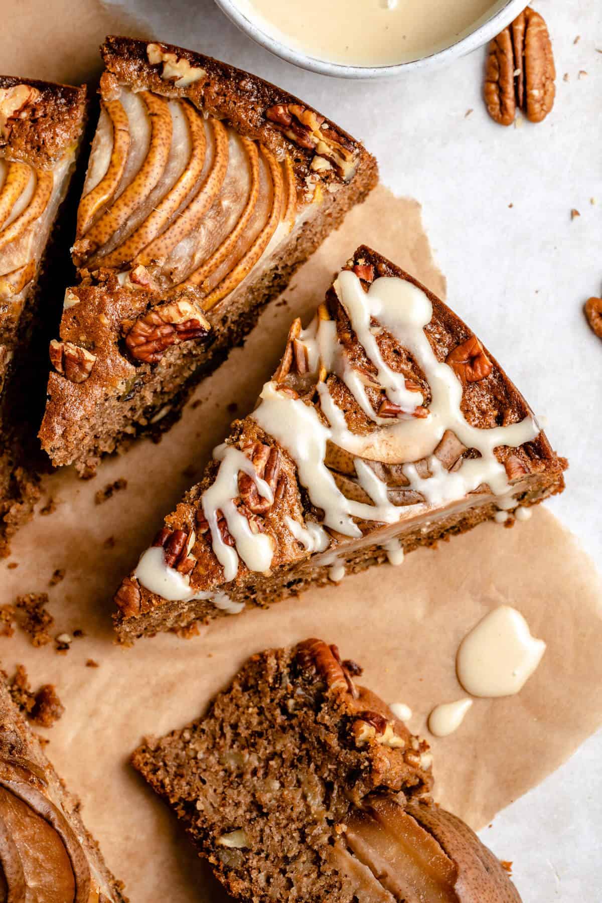 a close up image of a slice of cake drizzled with glaze, with more slices of cake and pecans around it