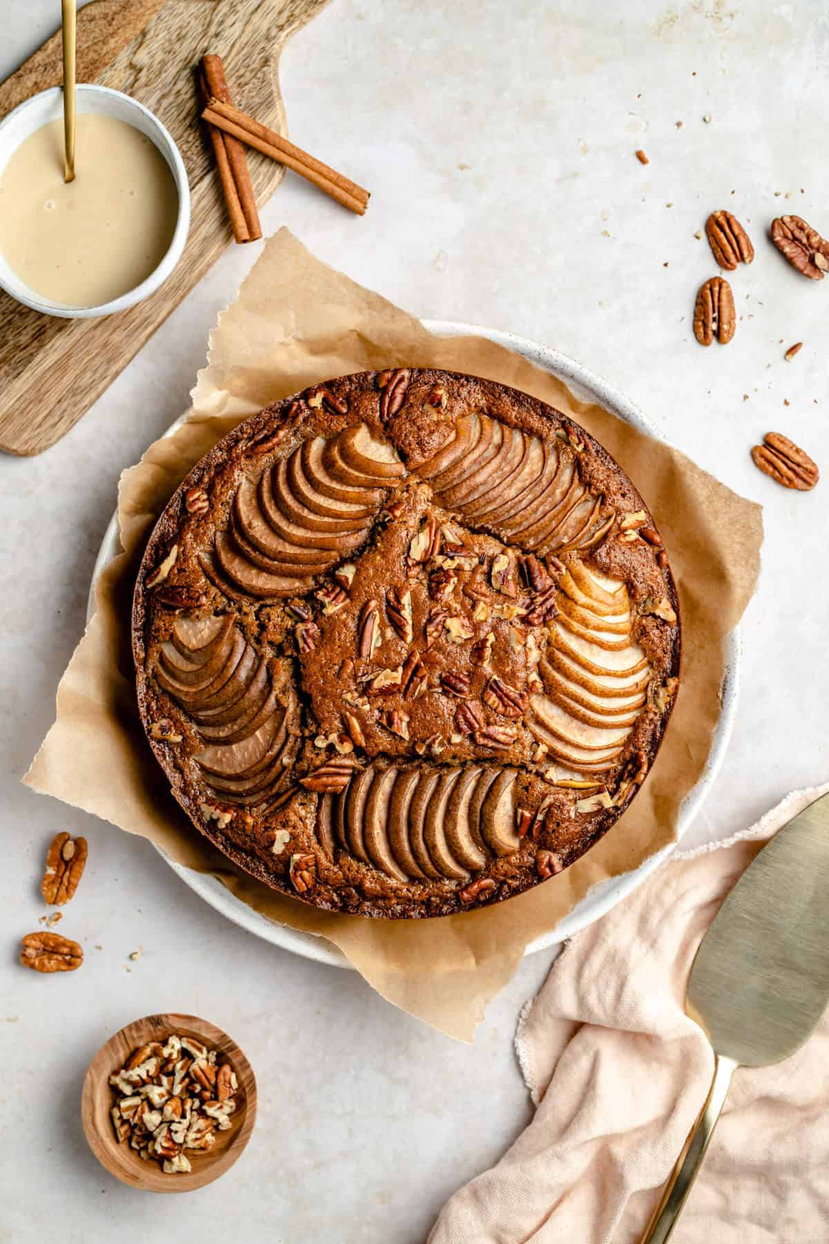 photo of the entire cake on a plate with parchment paper, surrounded by pecans, a bowl of tahini glaze and a gold cake server