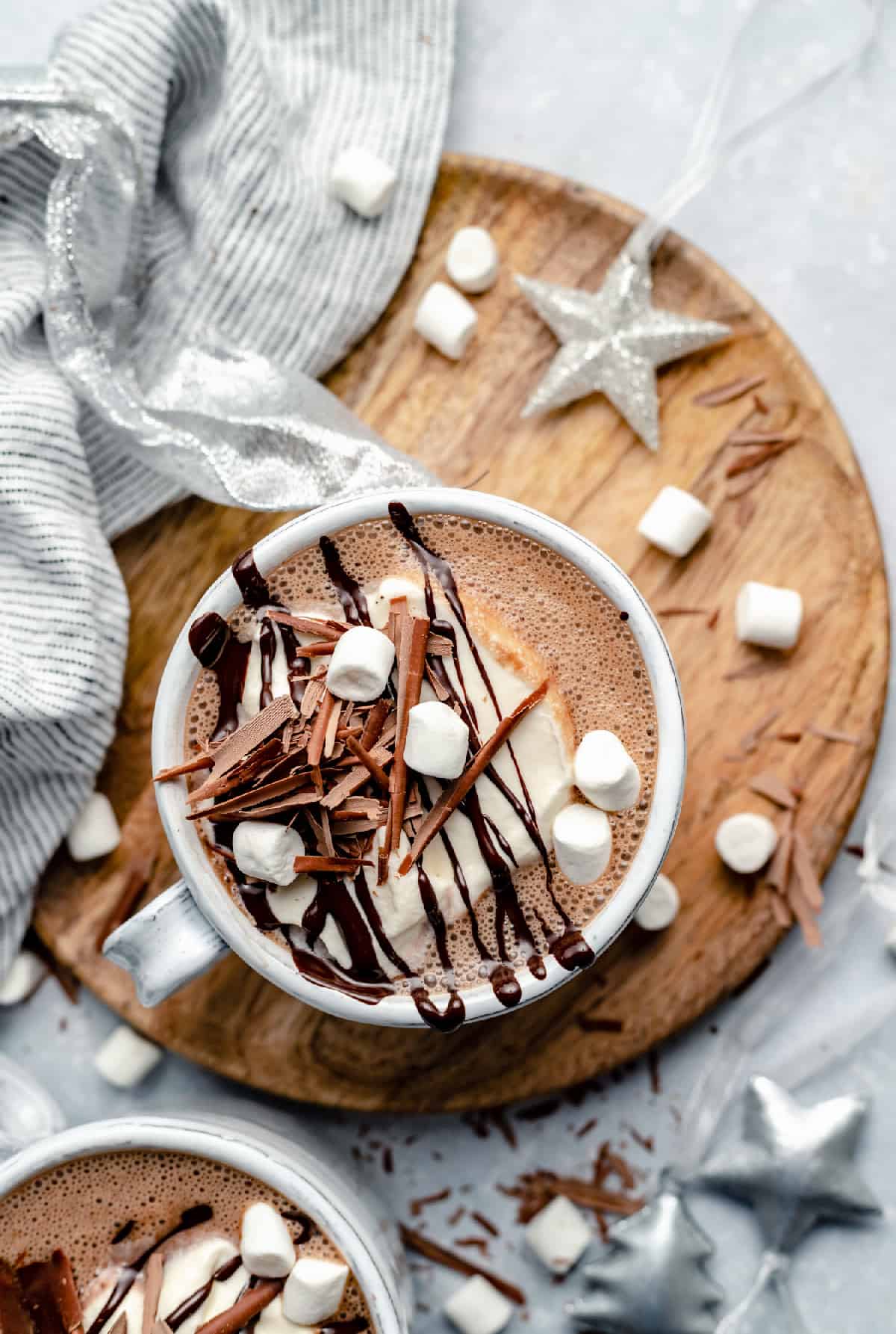 a mug of frothy peppermint hot chocolate topped with whipped cream, marshmallows and melted and shaved chocolate, on top of a wooden board and surrounded with silver christmas decorations