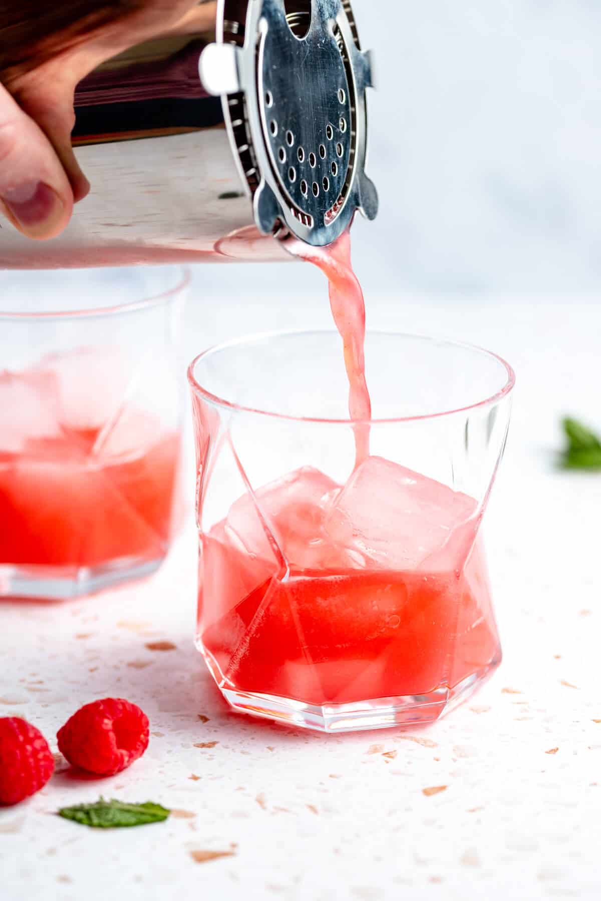 mocktail being poured over ice in a tumbler with raspberries and mint scattered around