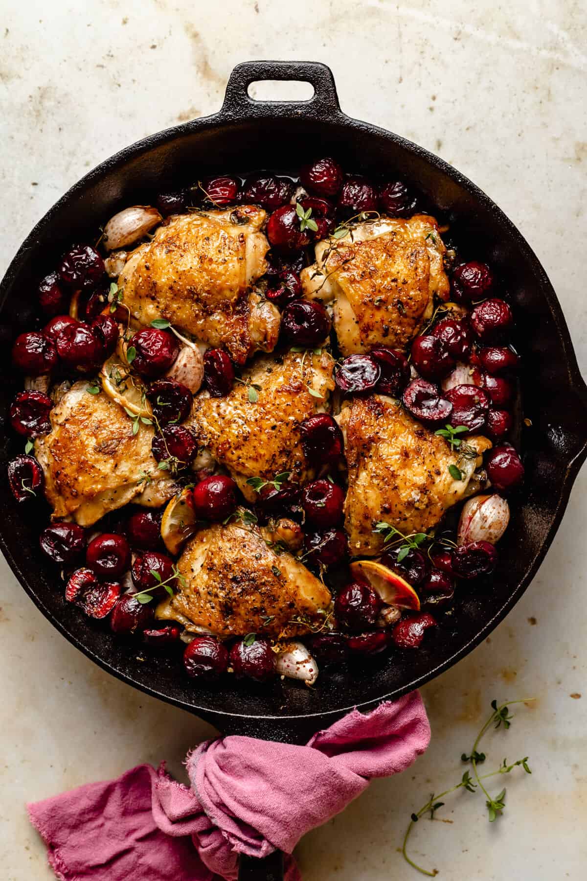 crispy chicken thighs in a skillet surrounded by juicy cherries with a pink napkin around the handle.