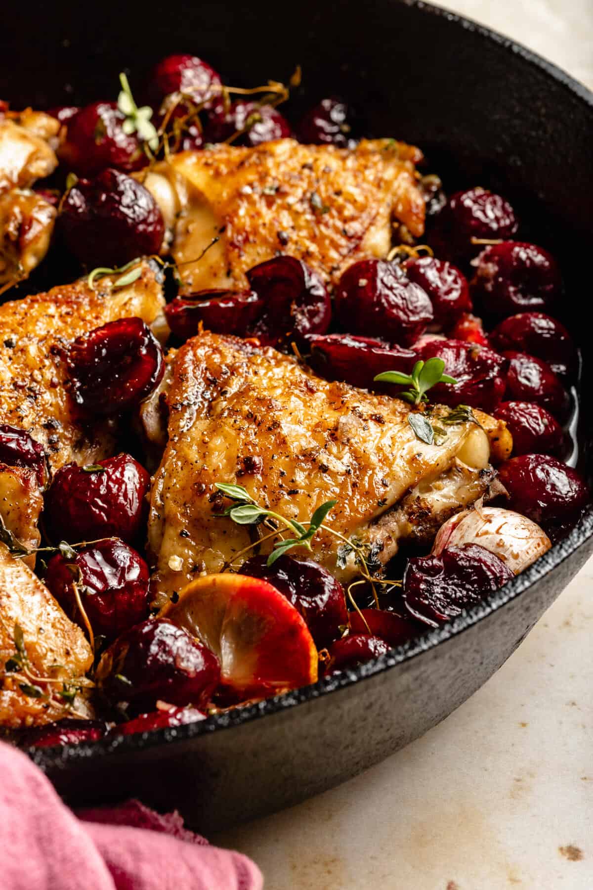 close up of crispy chicken thighs with cherries in a skillet with thyme leaves and lemon slices around.