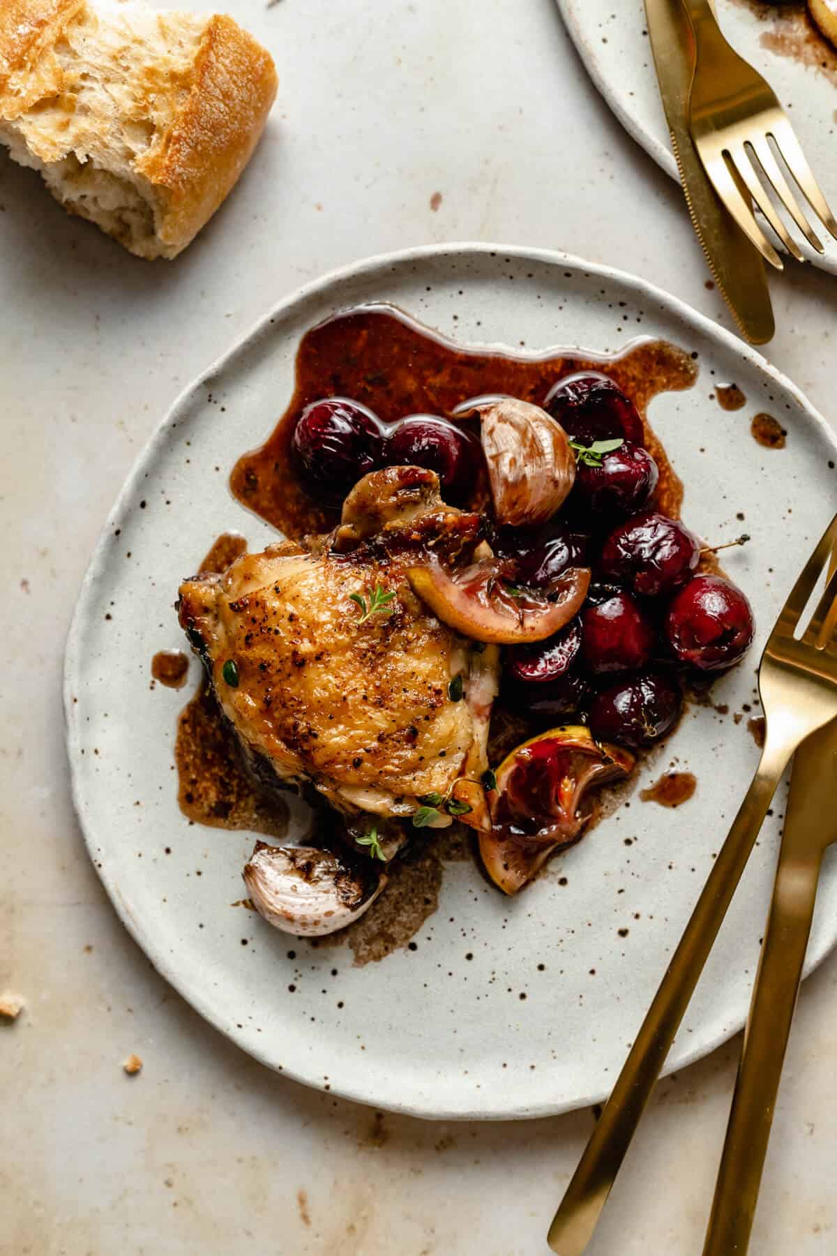 a crispy chicken thigh served on a plate with cherries, garlic and lemon with knife and fork to the side