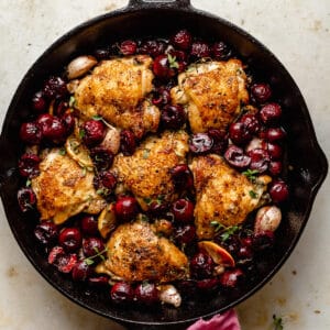 Cast iron chicken thighs with cherries in skillet with a pink napkin around the handle.