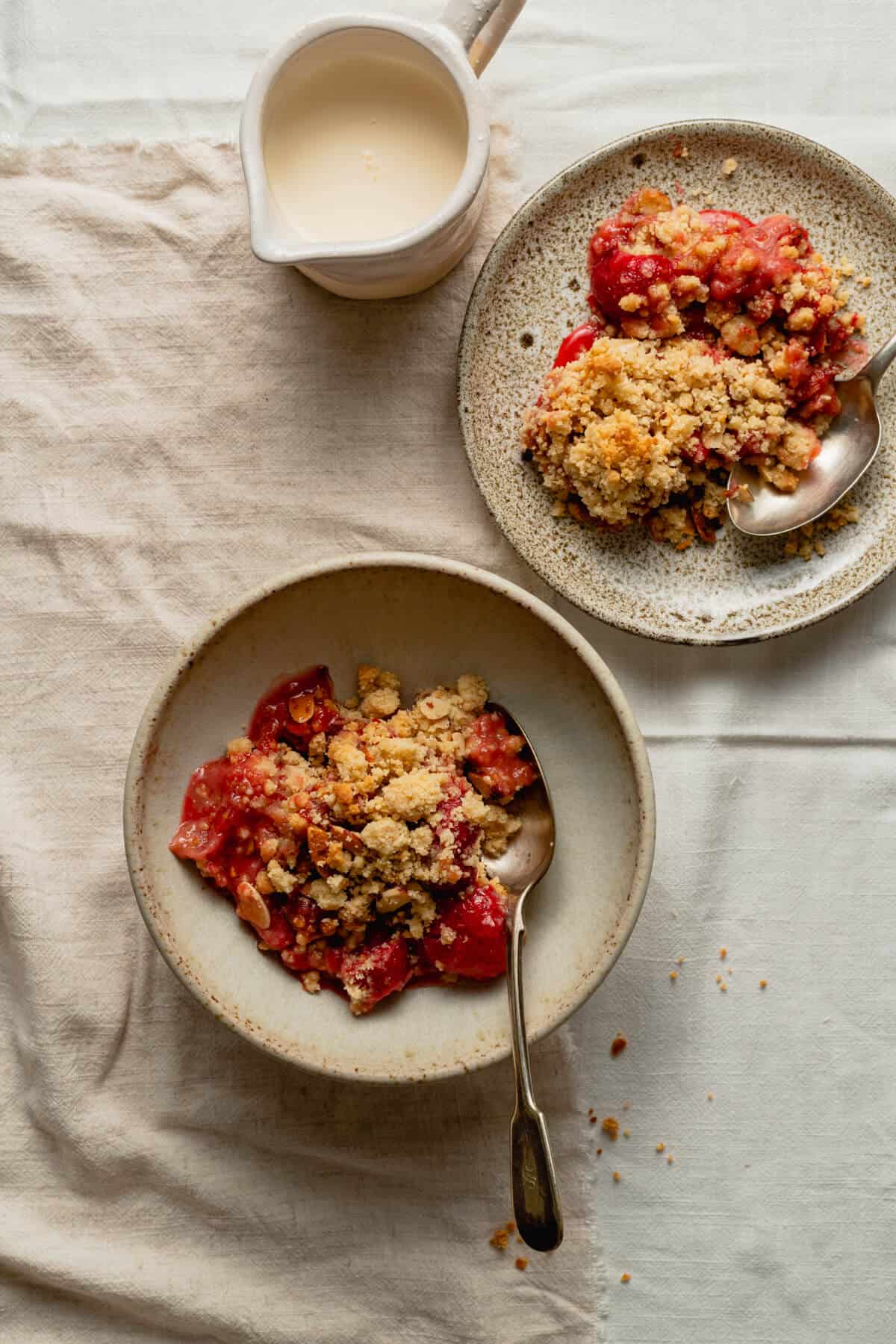 Strawberry crumble served up in a bowl and a plate with cream in a jug with spoons.