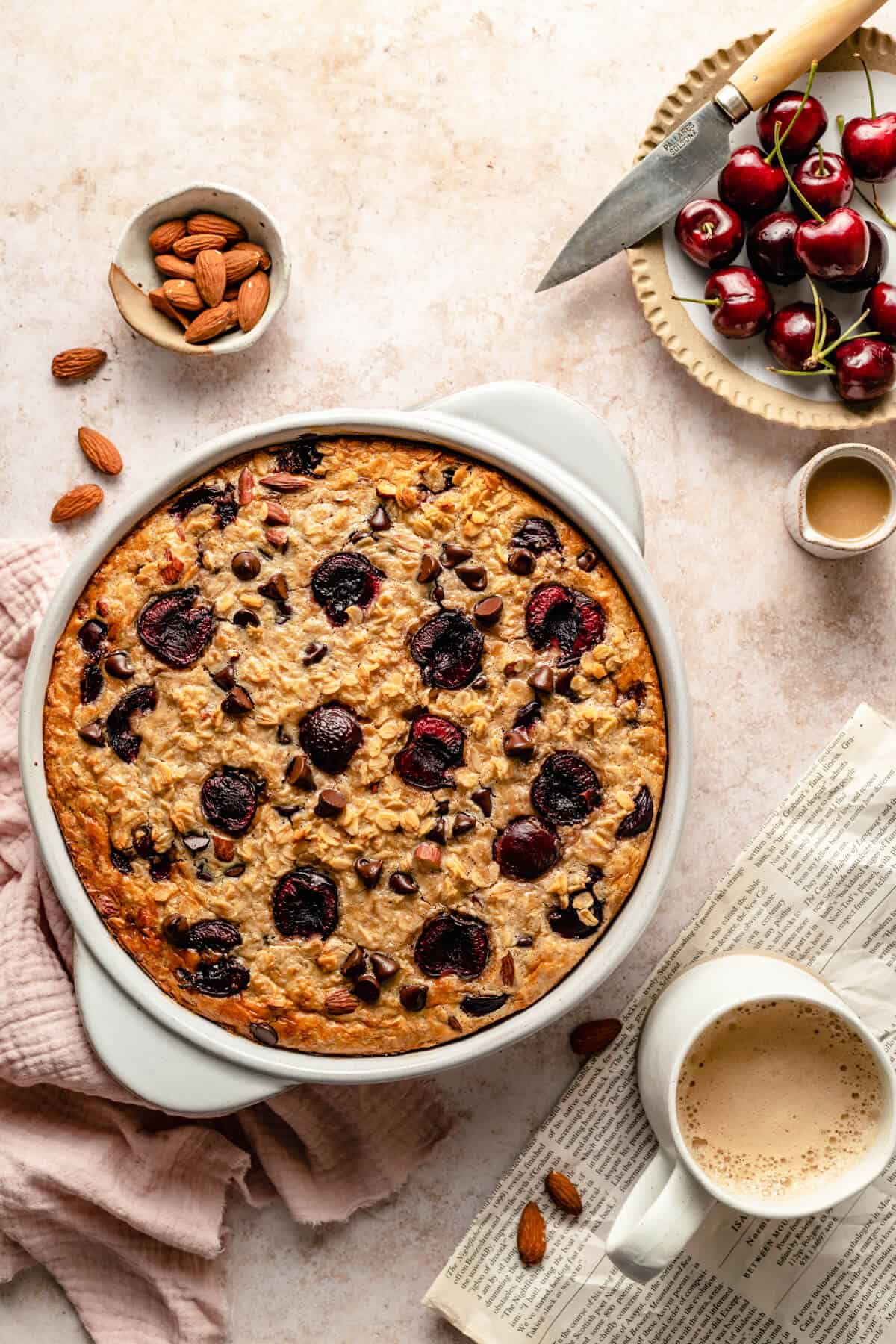 Cherry Chocolate Chip baked oatmeal in a baking dish with a mug of coffee and paper nearby.
