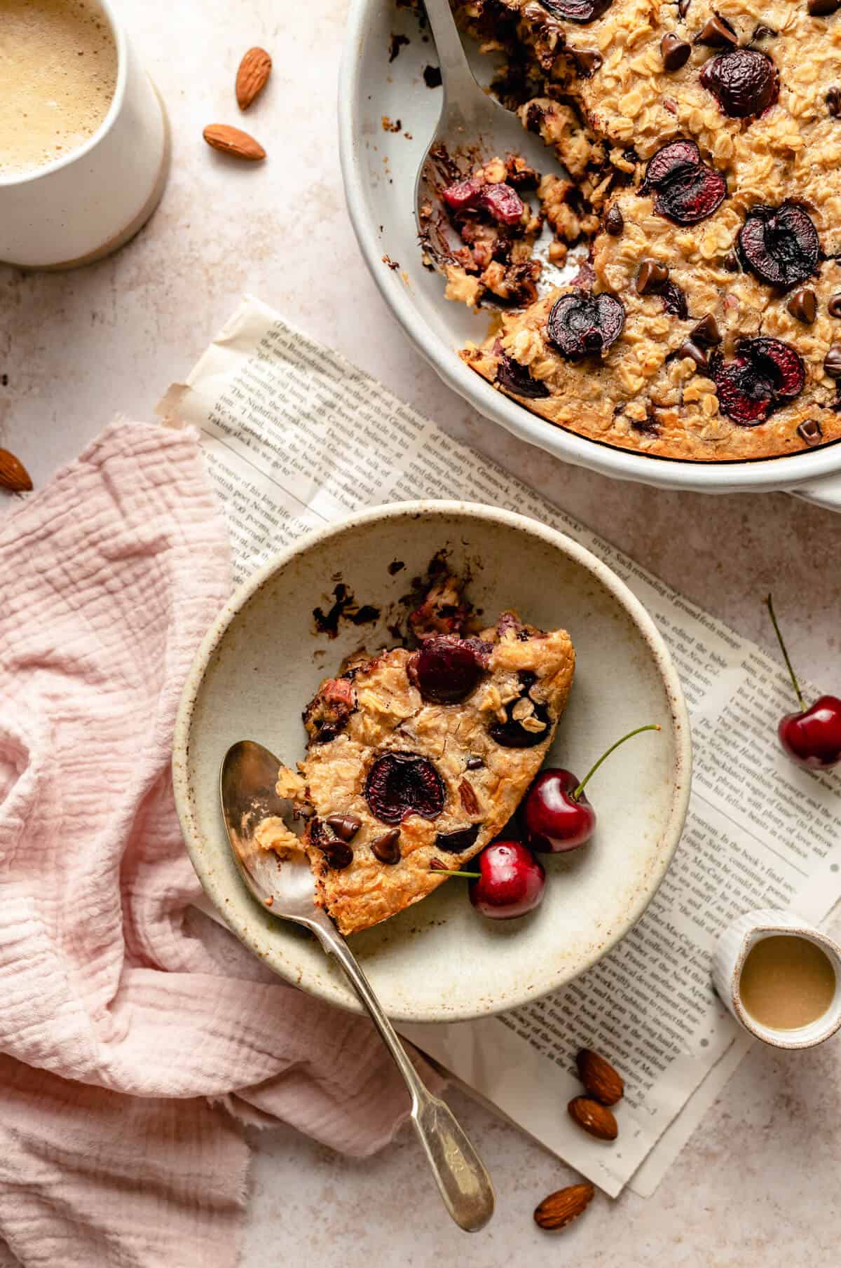 A serving of tahini cherry chocolate chip baked oatmeal in a bowl with a newspaper, cherries and a spoon. 