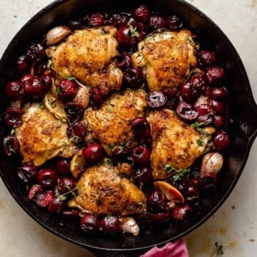 cast iron chicken thighs with cherries in a skillet with crispy skin and a napkin around the handle.