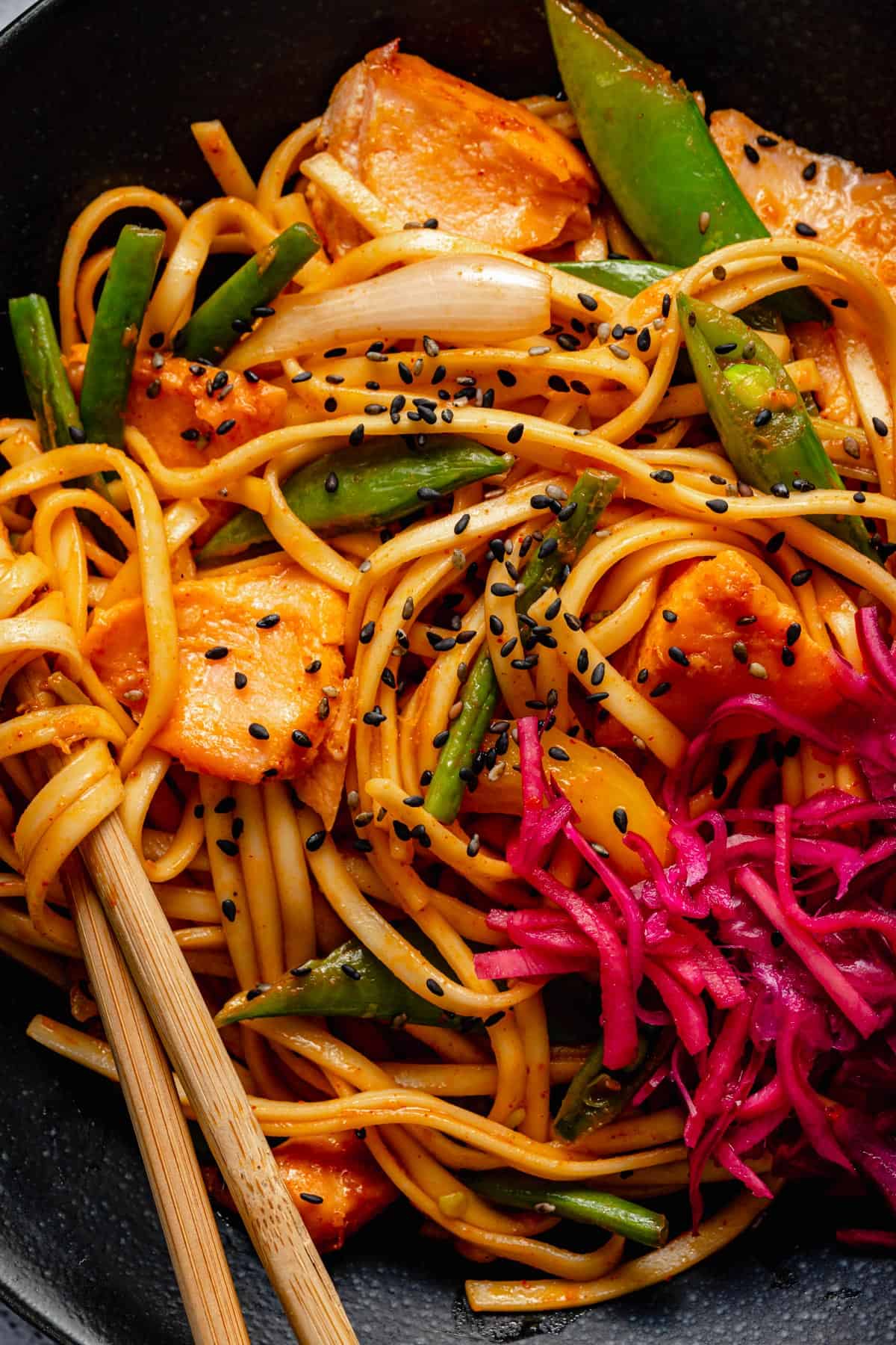 Close up of gochujang noodles and salmon in a bowl with chopsticks and pickle to the side.