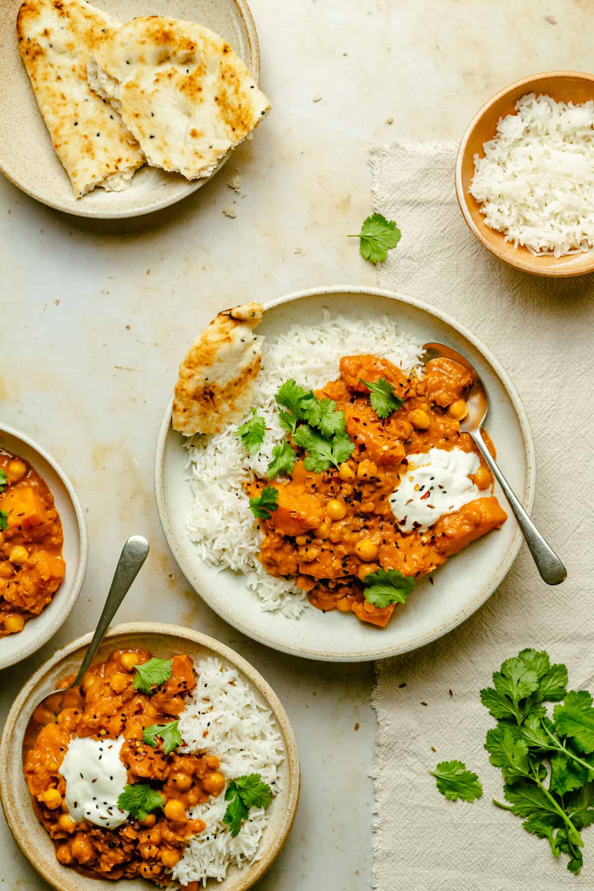 A couple of bowls of curry served with rice and yoghurt with spoons and coriander around.