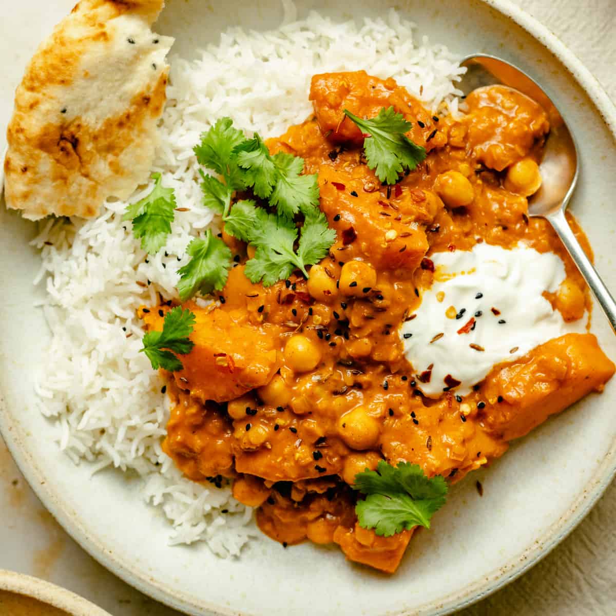 A bowl of butternut squash and chickpea curry with lentils on a bed of rice with a spoon and yoghurt
