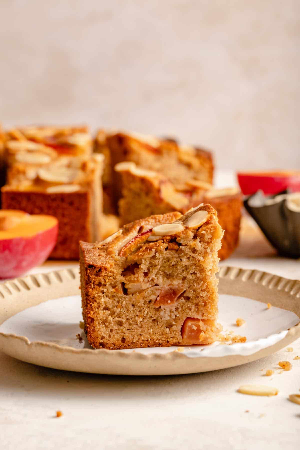 Slice of almond plum cake on a plate with crumbs, almonds, and a plum around it.