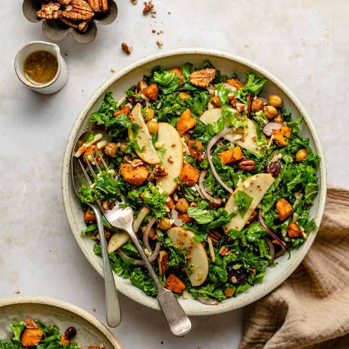 sweet potato kale salad in a serving bowl with a spoon and fork, dressing and pecans to the side