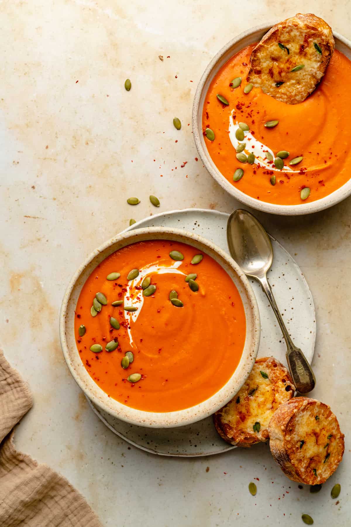 Two bowls of soup, topped with a swirl of yoghurt, pumpkin seeds and chilli flakes, with a spoon and some cheesy toasts on the side.