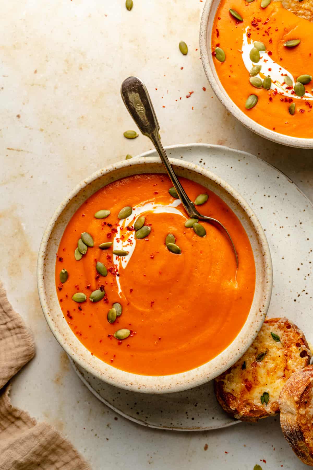 A bowl of butternut squash and red pepper soup, topped with yoghurt, pumpkin seeds and chilli flakes with a spoon in the bowl.