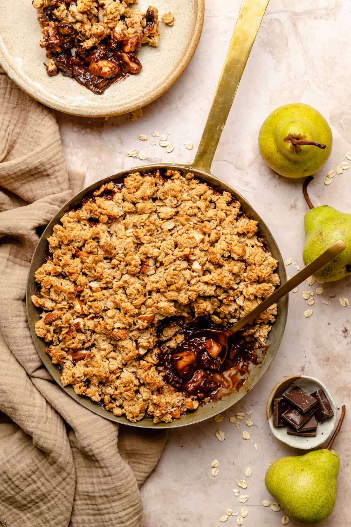 A copper pan with pear and chocolate crumble with a serving taken out of it and spoon showing the insides. Surrounded with pears, chocolate and oats.