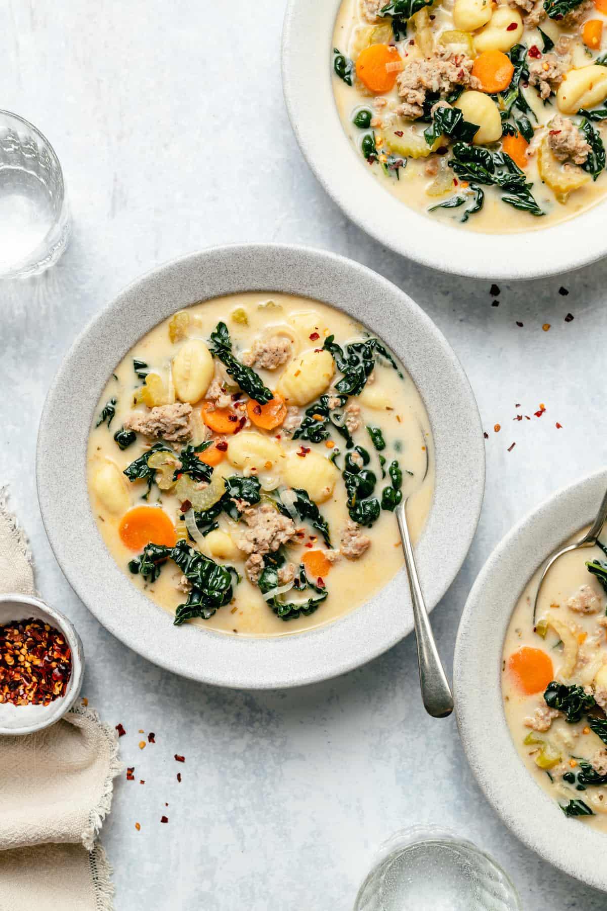 Bowls of sausage gnocchi soup with spoons, chilli flakes and glass of water on the side.