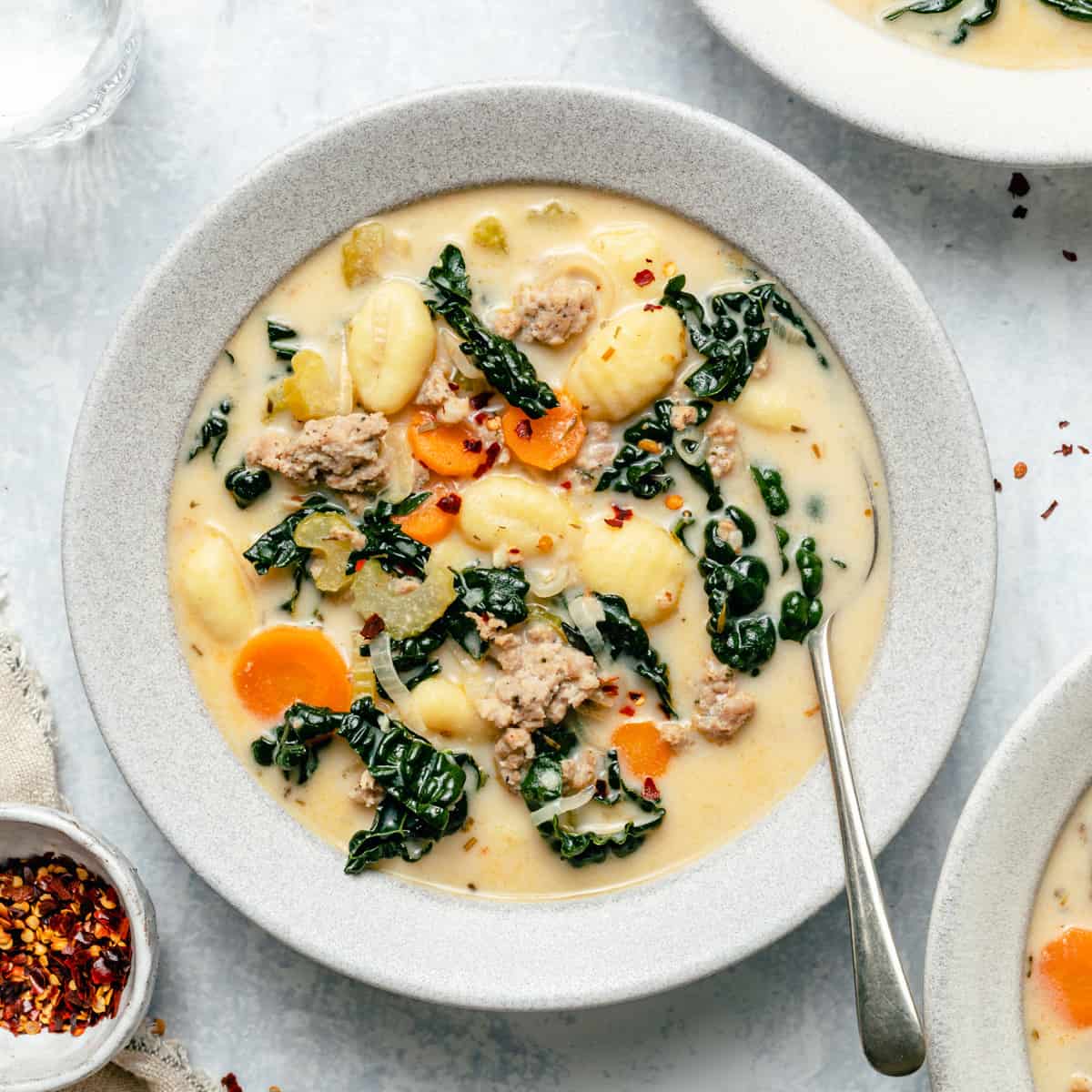 A bowl of sausage gnocchi soup with a spoon and chilli flakes on the side. 