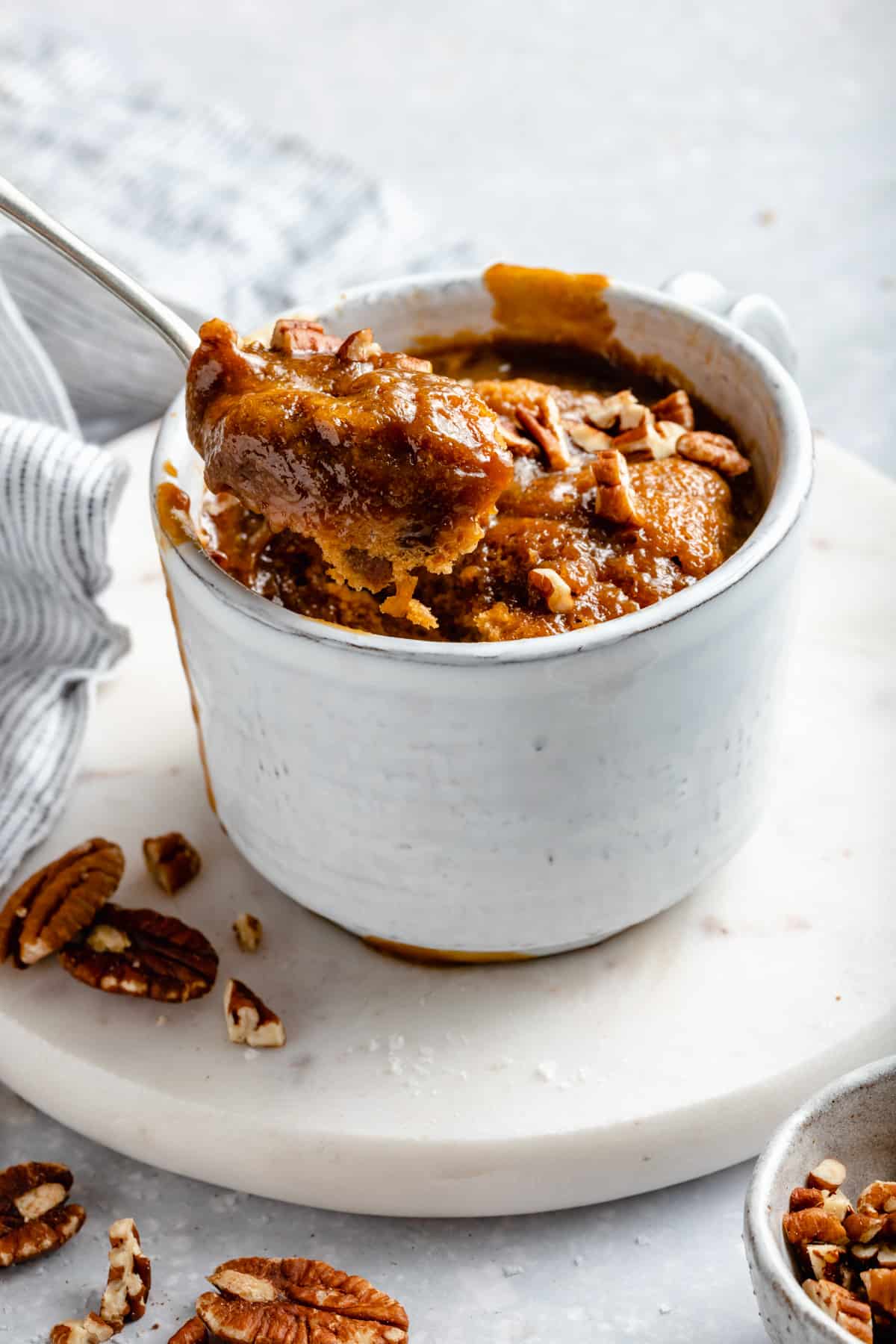 A spoon dipping into a sticky toffee mug cake with toffee sauce and nuts on top. 