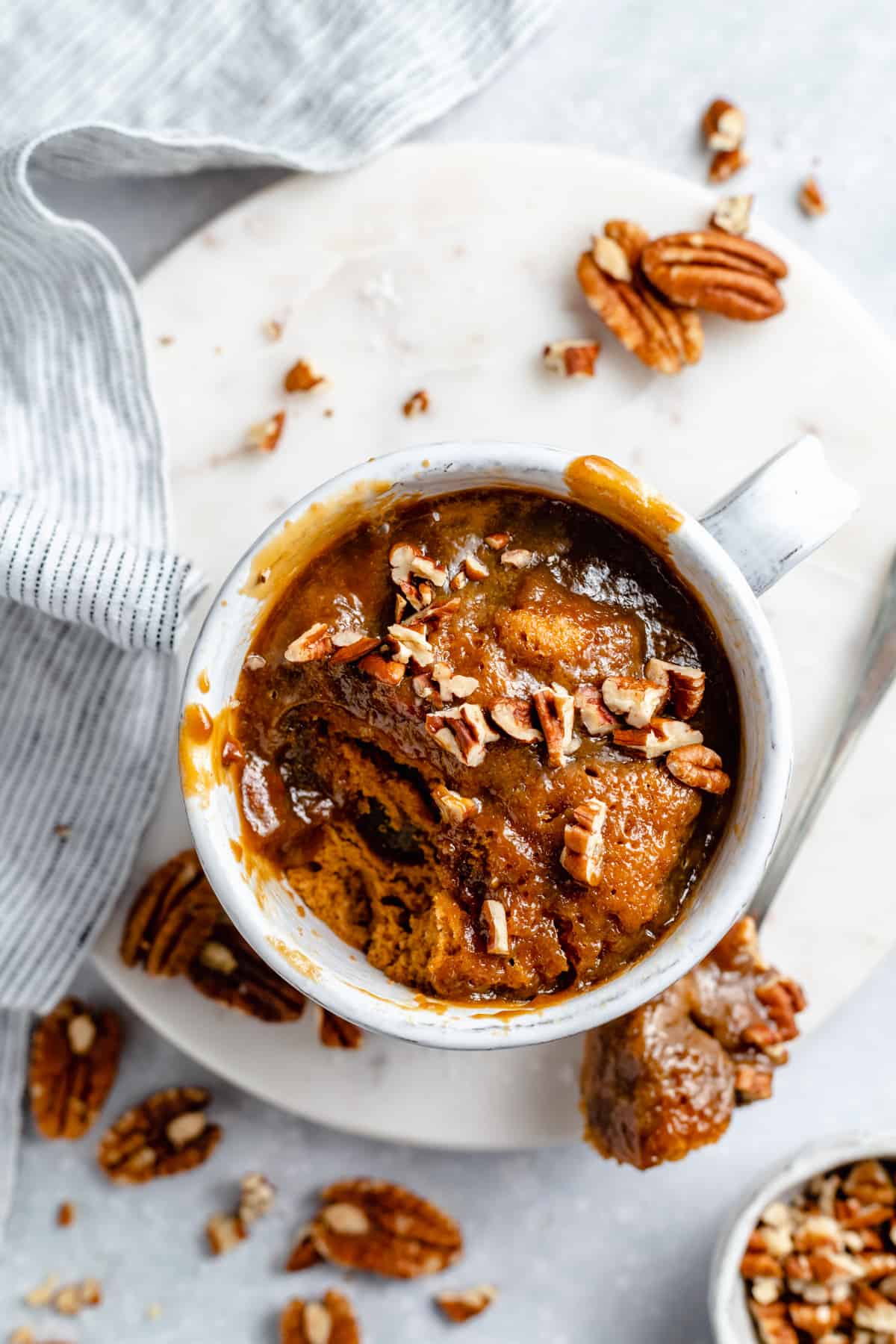 Microwave sticky toffee mug cake with a spoonful removed and the spoon on the side of a marble board with chopped nuts on top.