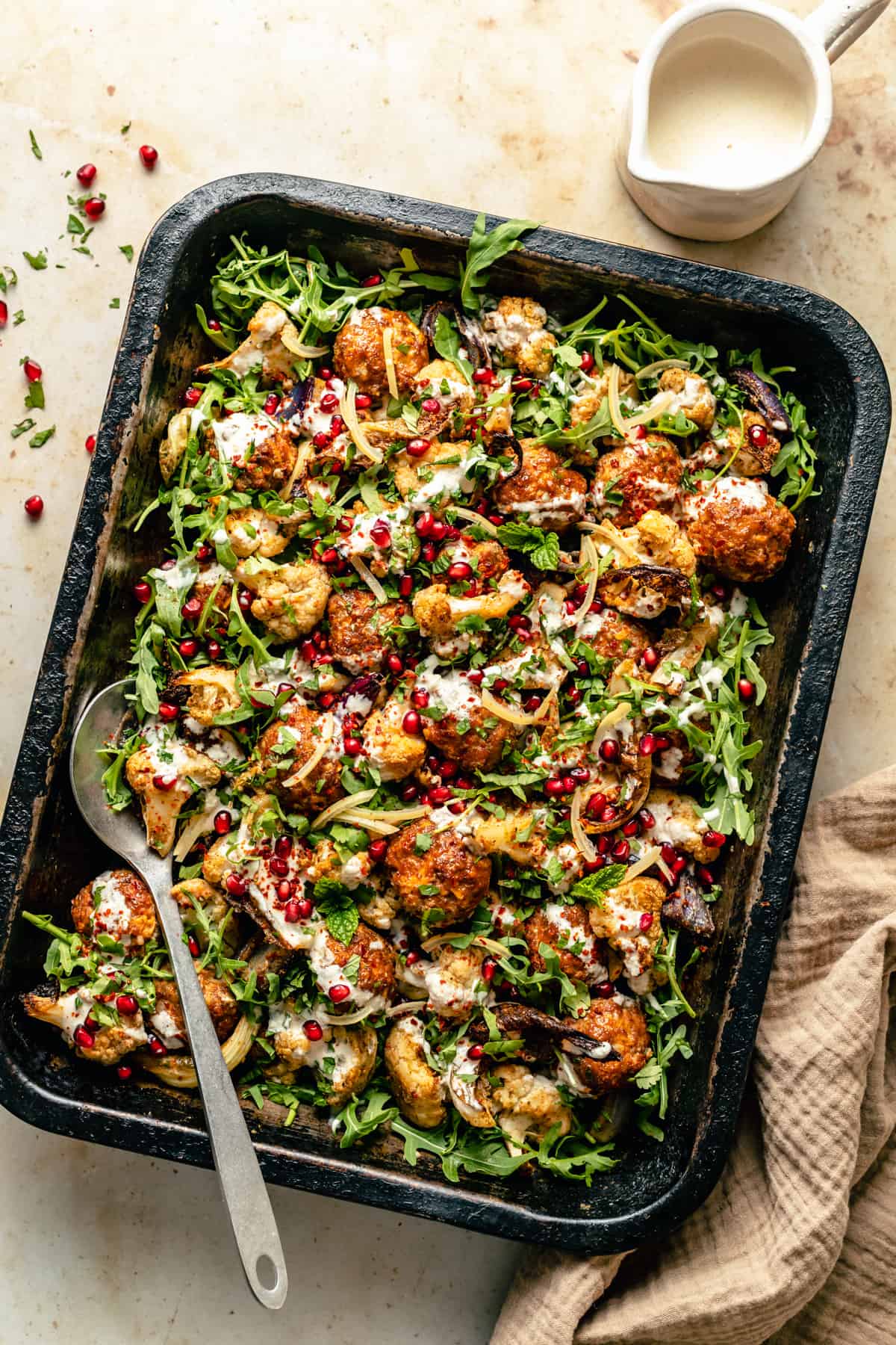 Baked spicy turkey meatballs on a bed of roasted cauliflower and arugula in a roasting tin dressed with lemon garlic yoghurt with a spoon.