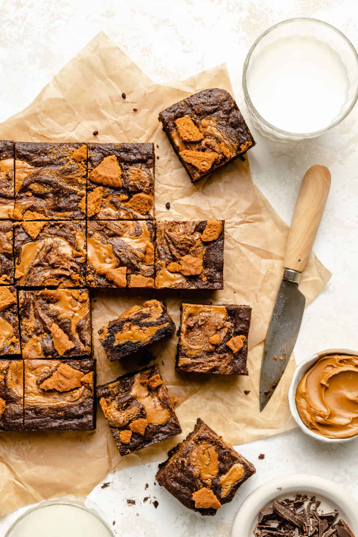 The biscoff brownies being cut up on parchment with a knife, bowl of biscoff and glass of milk to the side.