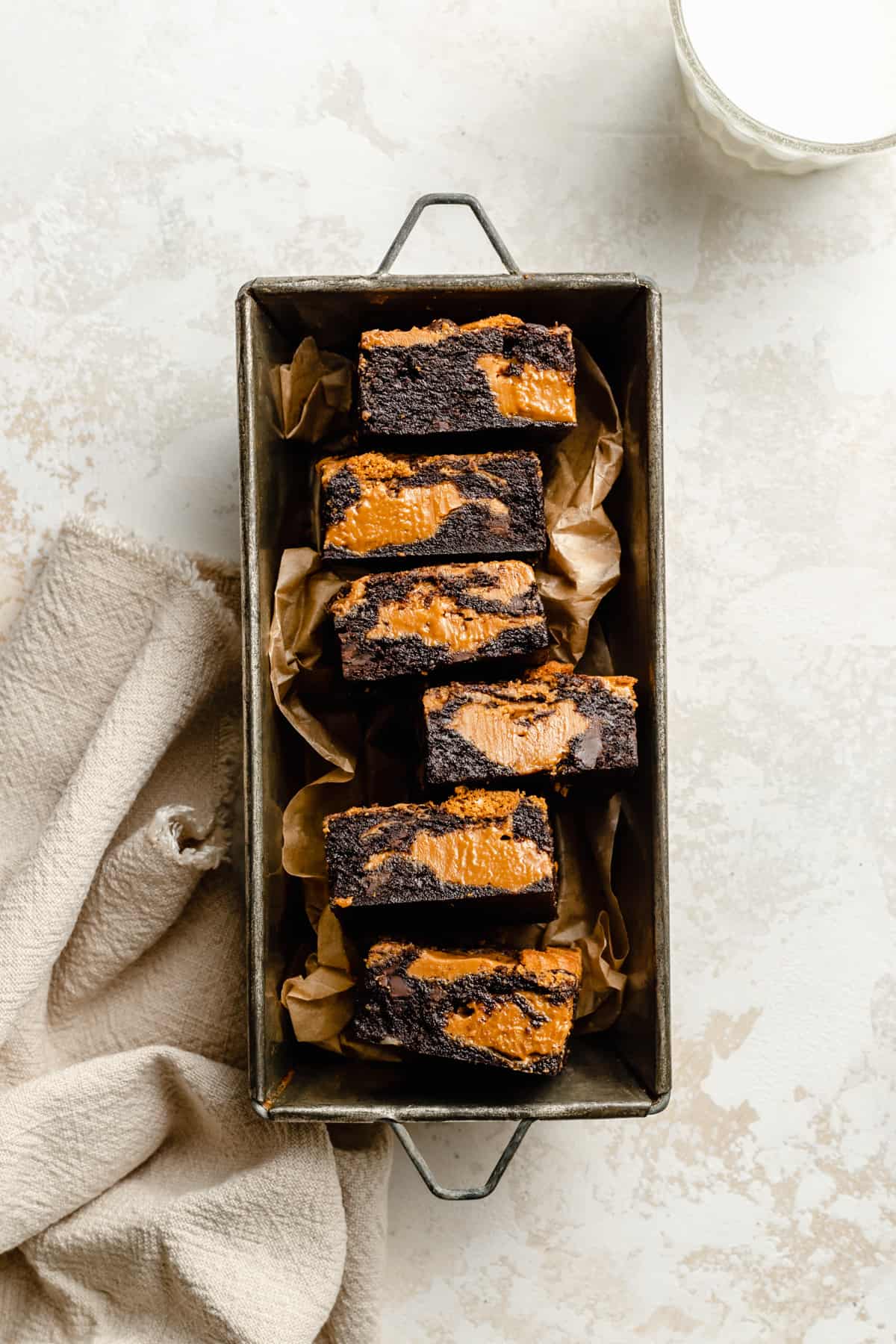An array of gooey fudgy chocolate biscoff brownies in a baking tin with a glass of milk to the side.