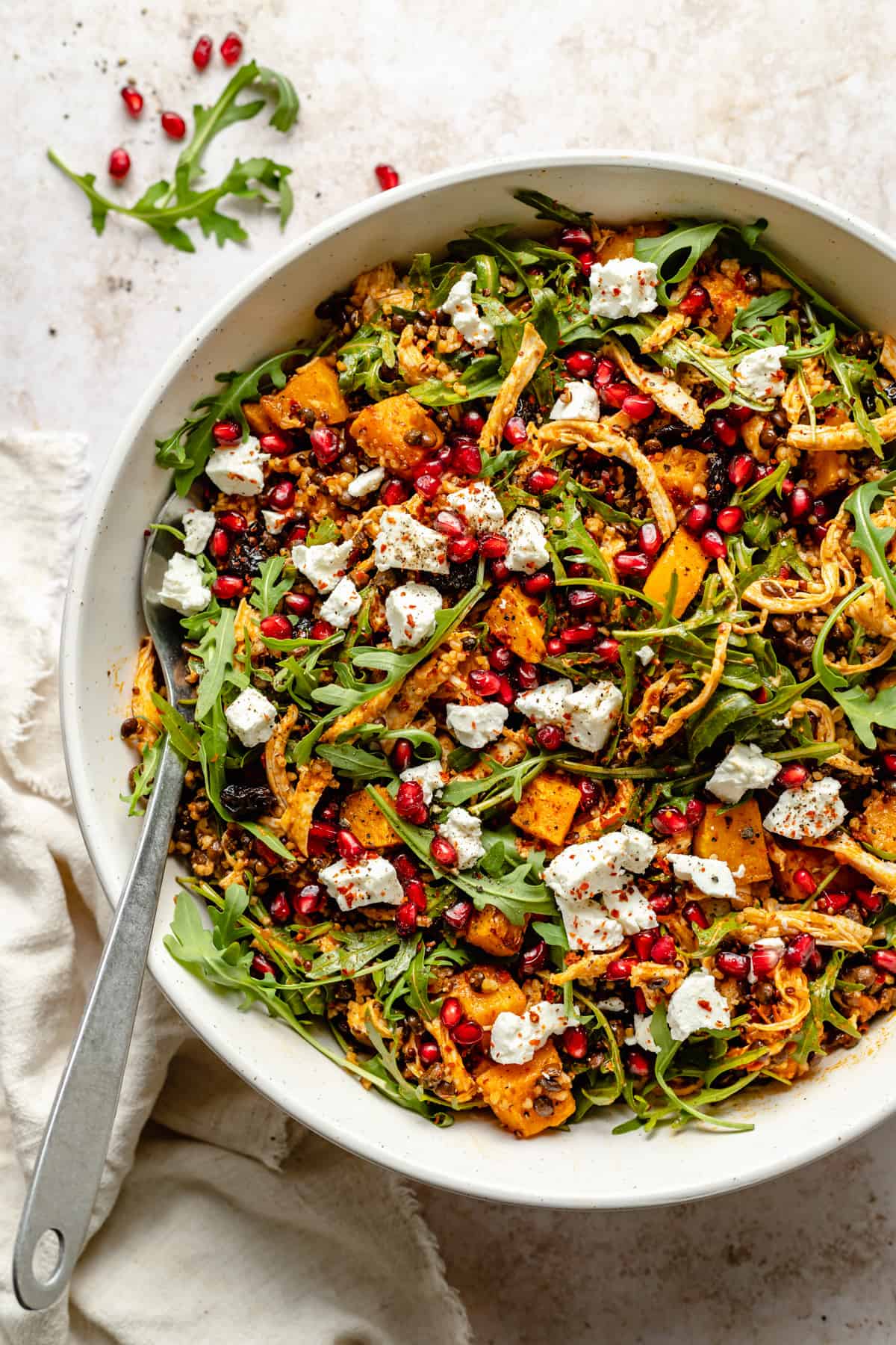 A large serving bowl with a dressed fall grain salad with a serving spoon, topped with feta and pomegranate arils.